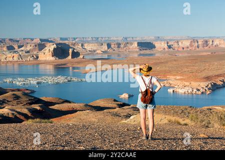 Rear view of woman walking at lakeshore looking at solitary rock formation Lone Rock in Wahweap Bay at Lake Powell, Glen Canyon Recreation Area, Page, Stock Photo