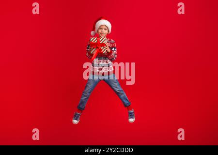 Full length photo of charming small boy jumping hold gift celebrate christmas dressed knitted sweater isolated on red color background Stock Photo