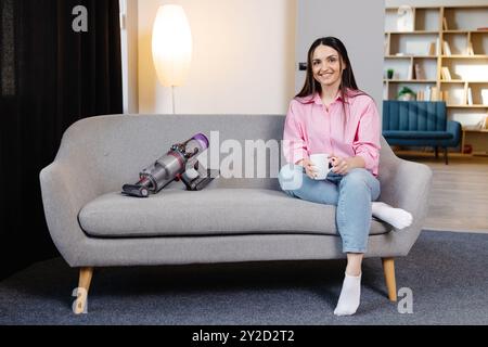 Young woman sitting on sofa next to cordless vacuum cleaner at home and holding cup in hands. Housework and chores concept. Cleaning appliances. Stock Photo