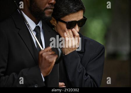 Medium shot of crying senior woman in black sunglasses wiping tears while clinging to husbands shoulder seeking comforting after grieving loss at memorial ceremony at gravesite, copy space Stock Photo