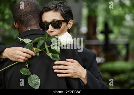 Medium shot of senior woman wearing black sunglasses grieving loss of loved one in comforting embrace of Black man at cemetery, copy space Stock Photo