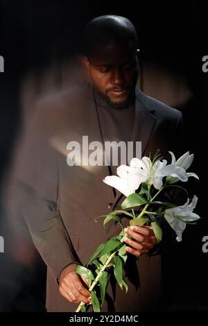 Vertical shot of pensive Black man in elegant suit holding delicate white lily illuminated by bright beam of spotlight in studio with black curtains, ghost glare effect Stock Photo