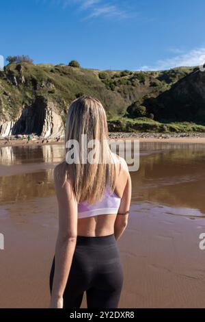 Woman with blonde hair walking on scenic beach in activewear, admiring the natural landscape and calm environment Stock Photo