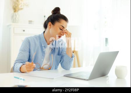 Photo of a tired arabian or indian business woman, company manager or digital seo, sits at a workplace, feels stressed and overworked, having migraine, headache, closes eyes and massaging bridgenose Stock Photo