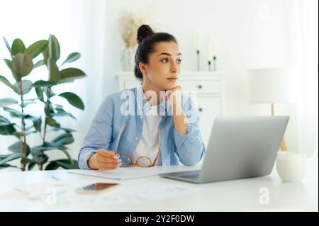 Worried business woman of arabian or indian ethnicity in blue a shirt, company manager, sits at a workplace with a laptop, feels overworked and stressed, looks away,thinking about deadlines Stock Photo