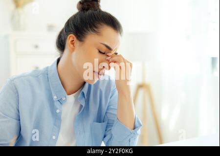 Close-up of a tired arabian or indian business woman, company manager or digital seo, sits at a workplace, feels stressed and overworked, having migraine, closes eyes and massaging bridgenose Stock Photo