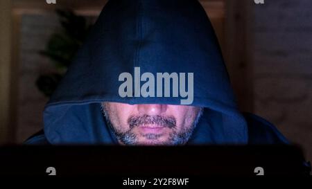 Bearded middle aged hooded hacker at laptop at night close up. Man using computer working from home Stock Photo