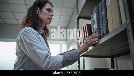 Female scientist or lab worker records voice message on mobile phone, uses AI voice chatbot and surfs the Internet while standing near shelf with folders. Modern archaeological scientific laboratory. Stock Photo