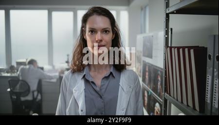 Modern archaeological science lab: Close up portrait of female scientist in lab coat smiling and looking at camera while standing near shelf with folders. Male colleague works in the background. Stock Photo