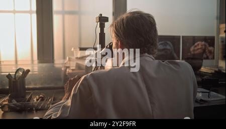 Mature chemist or microbiologist analyzes fossil sample under microscope. Scientist or archaeologist works with professional equipment in advanced archaeological science laboratory. Sunset light. Stock Photo