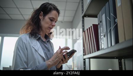 Female scientist or lab worker records voice message on mobile phone, uses AI voice chatbot and surfs the Internet while standing near shelf with folders. Modern archaeological scientific laboratory. Stock Photo