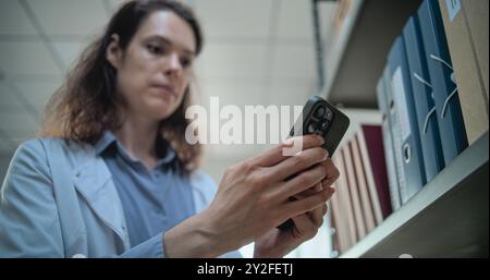 Close up of female scientist chatting, surfing the Internet on mobile phone, using AI chatbot for scientific research while standing near shelf with folders. Archaeological science lab. Low Angle. Stock Photo