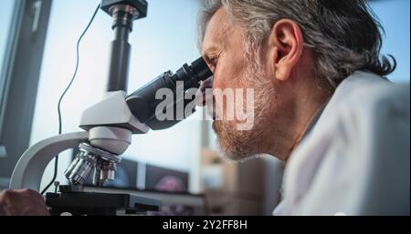 Close up of mature scientist conducting scientific research, analyzing fossil sample under microscope. Microbiologist works with advanced equipment in archaeological laboratory. Scientific method. Stock Photo