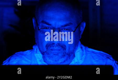 Bearded middle aged man working at laptop in blue light at night close up. Person in glasses using computer Stock Photo