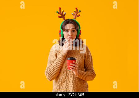 Worried young woman in reindeer horns with cup of coffee on yellow background Stock Photo