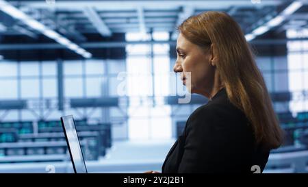 System administrator in data center typing on laptop, running error checking utilities to fix problems. Woman in server hub looking for network issues affecting rigs using notebook device, camera A Stock Photo