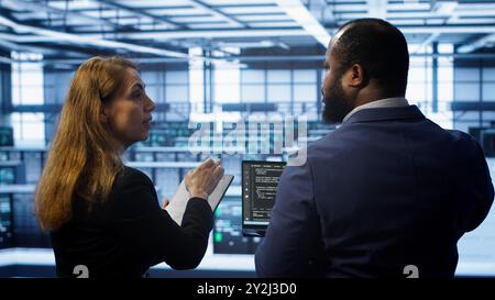 Teamworking programmers in data center overseeing server operations using laptop and tablet. Coworkers ensuring optimal performance and data integrity in server room using devices, camera A Stock Photo