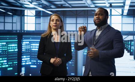 Teamworking technicians in high tech data center workspace looking at hologram visualization. Programmer and colleague working on securing server room hardware using AR tech, camera A Stock Photo