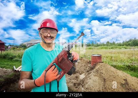 Mature smiling middle aged man in construction helmet and safety glasses holds corded hammer drill with chisel in his hands. Stock Photo