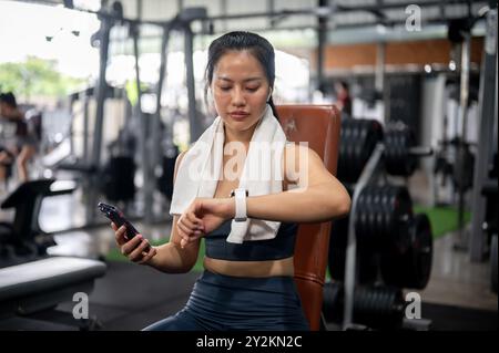 A sporty Asian woman in athletic wear is tracking her burnt calories, checking her workout results on her smartwatch while resting on a bench press at Stock Photo
