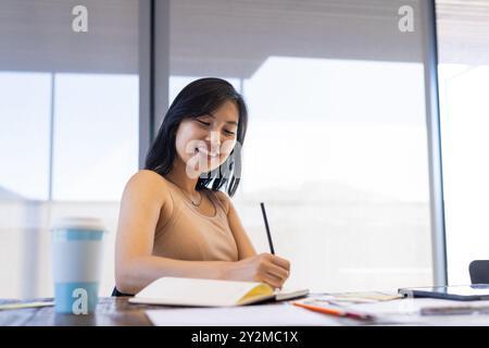 At office, Smiling asian woman writing in notebook at desk with coffee cup and papers Stock Photo