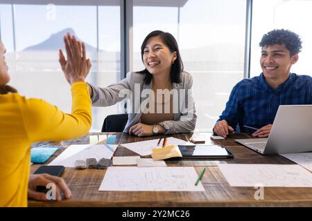 At office, Collaborating on project, diverse team high-fiving with laptop and sketches Stock Photo