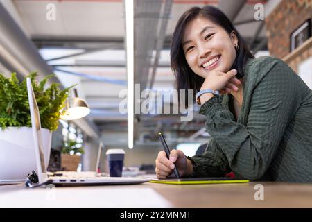 Smiling asian woman using digital pen and tablet while working in modern office Stock Photo