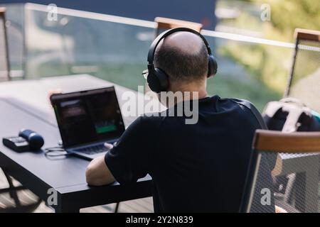 Back view from professional video editor editing video at vacation. Middle aged man using laptop and hard drives. Wireless headphones  Stock Photo
