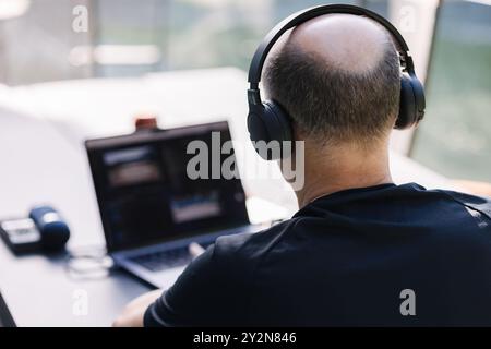 Back view from professional video editor editing video at vacation. Middle aged man using laptop and hard drives. Wireless headphones  Stock Photo