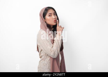 Young Asian Muslim woman wearing headscarf veil hijab looking to camera while putting a finger in front of lips gesturing stay silent or stay quiet, i Stock Photo