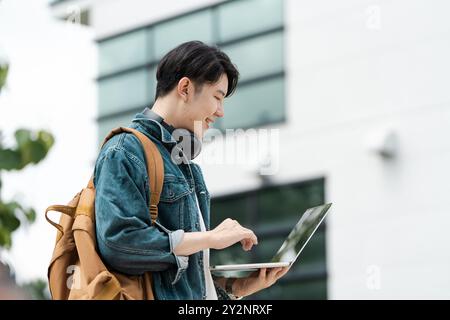 Young Man Using Laptop Outdoors with Modern Building in Background, Wearing Denim Jacket and Backpack Stock Photo