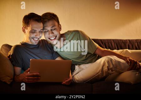Two men share laughter and joy while using a laptop, embracing on the couch. Stock Photo