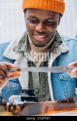 Smiling African-American man in beanie and denim jacket examines negatives of film, taking photographs and developing analog film. Photographer examines pics developed tape, rejoices result shooting. Stock Photo