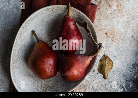 Three ripe organic pears with a reddish hue resting in a textured ceramic bowl, adorned by dry leaves, set against a gritty surface Stock Photo