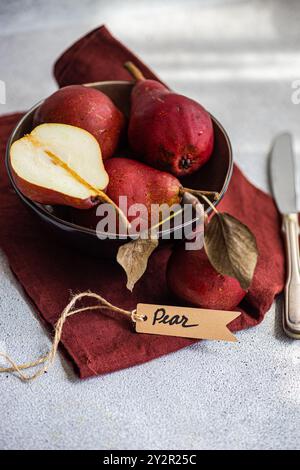 A collection of deep red organic pears, one sliced into a half, neatly placed in a dark metal bowl that rests on a burgundy cloth adorned with a paper Stock Photo