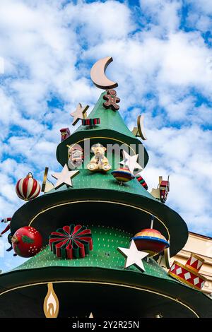 Low angle of a whimsical Christmas tree decoration towers against a blue sky with clouds in Tbilisi, Georgia, featuring colorful ornaments and a teddy Stock Photo