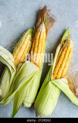 Three fresh corn cobs with partially peeled husks placed on a rustic surface The vibrant yellow kernels and green husks highlight the natural freshnes Stock Photo