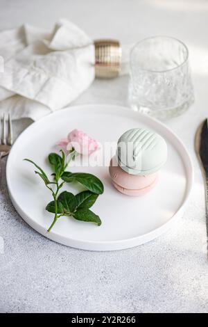 A delicate display of pastel macarons stacked atop each other beside a soft pink rose on a pristine white dining plate, accented with a clear glass an Stock Photo