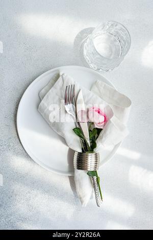 An elegant table setting featuring a pristine white plate, a folded napkin with cutlery, and a pink rose in a silver holder, alongside a clear glass o Stock Photo