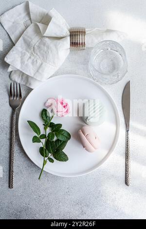 A refined dining setup showcasing a white plate adorned with a fresh rose and pastel-colored macarons The table is elegantly completed with a folded c Stock Photo