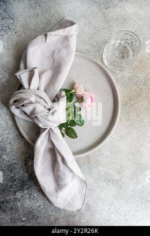 A minimalist composition featuring delicate pink roses and a knot of gray linen napkin, set on a textured ceramic plate with a clear glass in the corn Stock Photo