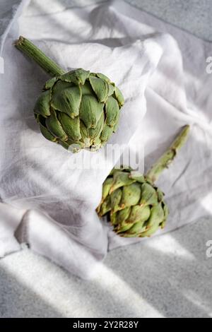 High-quality image of two raw artichokes placed on a crumpled white linen cloth, emphasized by soft natural lighting Ideal for food and nutrition them Stock Photo