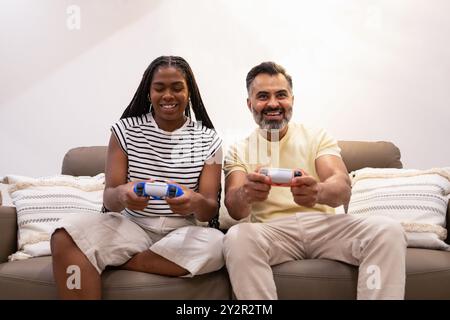 A joyful Black woman and an indian man enjoy a fun gaming session on a cozy sofa, showcasing a relaxed and cheerful domestic scene Stock Photo