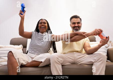Joyful couple sharing a fun moment, laughing and playing with handheld game controllers on their couch, radiating happiness and enjoyment in their coz Stock Photo