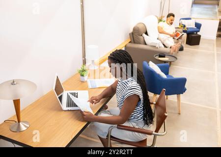A black woman works on her laptop at a minimalist styled desk while an indian man reads in the background, reflecting a peaceful and modern home setti Stock Photo