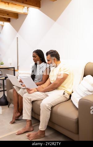 A multiethnic couple is sitting on a sofa, working together on a laptop in a cozy, well-lit living room, showcasing a relaxed and collaborative enviro Stock Photo