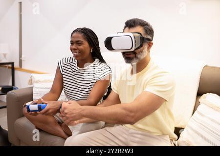 A cheerful Black woman and a man wearing a VR headset play a video game together in a cozy living room, laughing and enjoying a casual, fun moment at Stock Photo