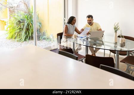 A cheerful indian man and black woman discuss documents at a modern glass dining table, with a bright garden view in the background Stock Photo