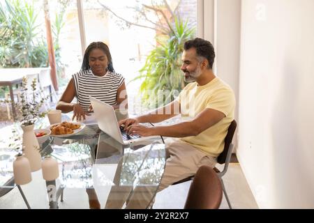 A black woman enjoys breakfast while an indian man works on his laptop in a bright home setting, enhancing productivity and comfort Stock Photo
