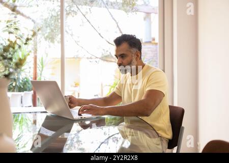Focused middle-aged indian man using a laptop at a glass table, illuminated by natural light from nearby window, in a calm home setting Stock Photo